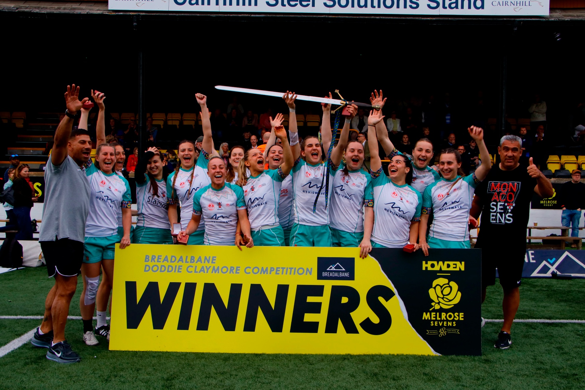 Sevens squad cheering behind a winners banner with the ceremonial sword
